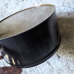 May include: A black metal container with a smooth, shiny surface. The container is cylindrical and has a slightly rounded top.