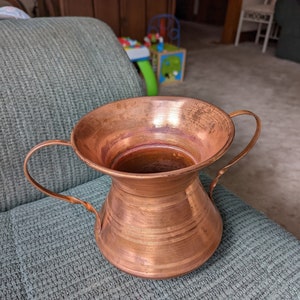 May include: A copper vase with two handles. The vase has a wide mouth and a narrow base.
