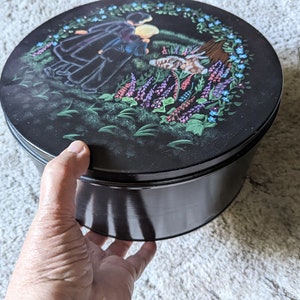 May include: A black round tin with a painted scene of two people in a field of flowers. The lid of the tin has a floral design with a wreath of flowers around the edge.
