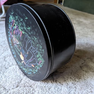 May include: Black round tin with a floral design featuring purple flowers and a couple of figures in a forest setting.