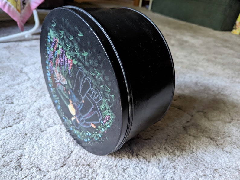 May include: Black round tin with a floral design featuring purple flowers and a couple of figures in a forest setting.