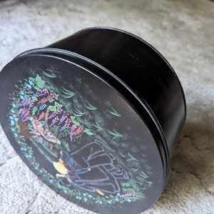 May include: Black round tin with a floral design featuring a girl in a black dress and a cat. The design includes purple, pink, and blue flowers.