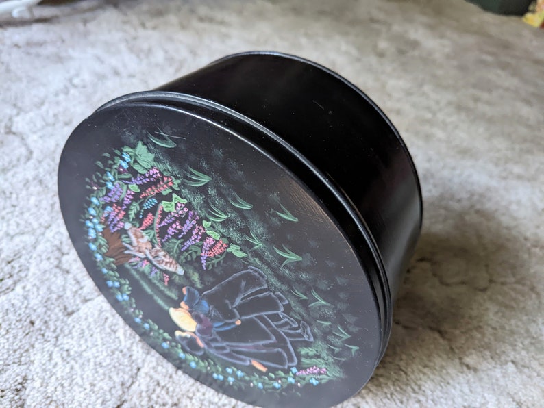 May include: Black round tin with a floral design featuring a girl in a black dress and a cat. The design includes purple, pink, and blue flowers.
