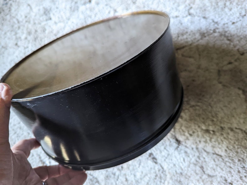 May include: A black metal container with a smooth, shiny surface. The container is cylindrical and has a slightly rounded top.