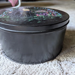 May include: A black round tin with a painted design of two figures in a garden with flowers. The tin has a lid and is closed.