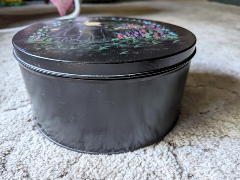 May include: A black round tin with a painted design of two figures in a garden with flowers. The tin has a lid and is closed.
