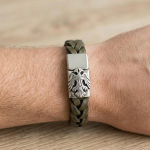 May include: Olive green braided leather bracelet with a silver-tone clasp featuring a detailed eagle emblem. The bracelet is shown on a wrist, showcasing the texture and design of the accessory.