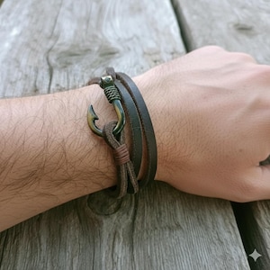 May include: A brown leather bracelet with a bronze-colored fish hook clasp. The bracelet wraps around the wrist and features a braided cord detail. The hook is a prominent design element.