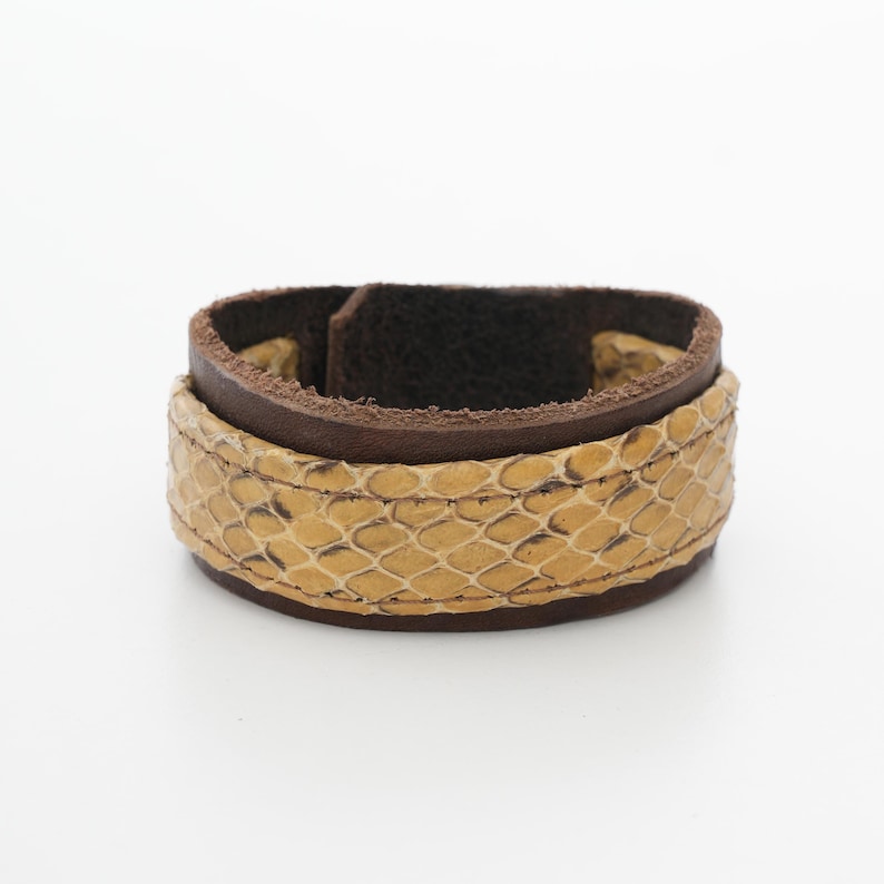May include: A wide leather bracelet with a snakeskin pattern in tan and brown. The bracelet has a dark brown leather base and a lighter tan snakeskin overlay. The bracelet is approximately 2 inches wide.