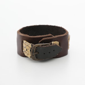 May include: A brown leather cuff bracelet with a dark brown interior. The bracelet features a snakeskin accent and a dark metal buckle. The cuff is approximately 2 inches wide.