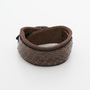 May include: A brown leather cuff bracelet with a textured, snakeskin-like pattern. The bracelet features a buckle closure and is designed to wrap around the wrist. The leather appears to be a rich, dark brown color.