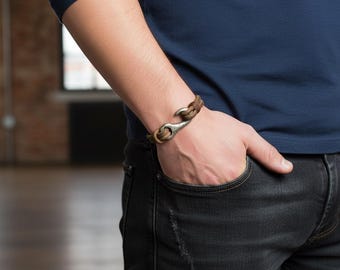 May include: A brown leather bracelet with a silver-colored hook clasp. The bracelet is made of braided leather and is worn on a wrist. The style is casual and rugged, suitable for everyday wear. The bracelet is a stylish accessory.