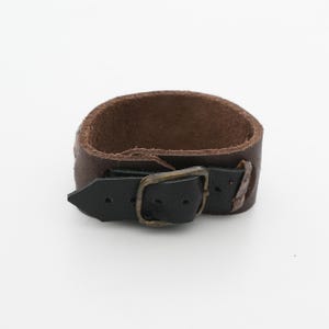 May include: A brown leather cuff bracelet with a black leather strap and a brass-colored buckle. The bracelet is approximately 1.5 inches wide and has a rugged, worn appearance.
