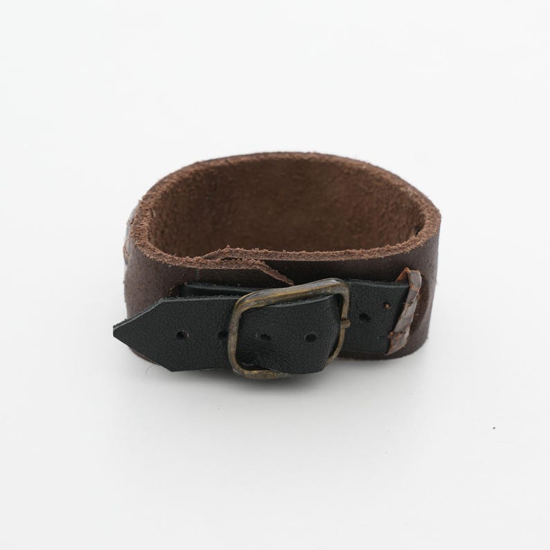 May include: A brown leather cuff bracelet with a black leather strap and a brass-colored buckle. The bracelet is approximately 1.5 inches wide and has a rugged, worn appearance.