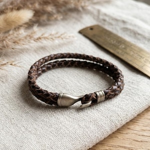 Puede incluir: Una pulsera de cuero marrón con un diseño trenzado. La pulsera presenta un cierre de color plateado. La pulsera se muestra sobre una superficie de lino de color neutro, con una regla de latón en el fondo.