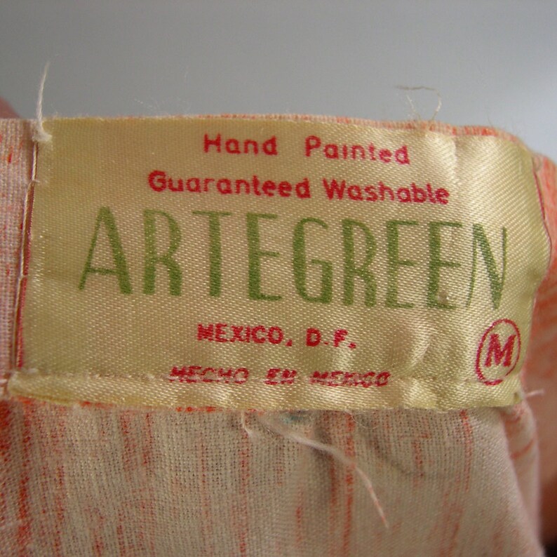 Pu&ograve; includere: Un'etichetta gialla con il testo "Hand Painted Guaranteed Washable ARTEGREEN MEXICO, D.F. HECHO EN MEXICO" in rosso e verde. L'etichetta ha anche una "M" cerchiata in rosso.