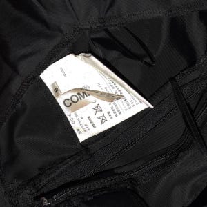 May include: Black clothing label with white text that reads "COMME des GARCONS" and "S" for size. The label also includes Japanese text and washing instructions.