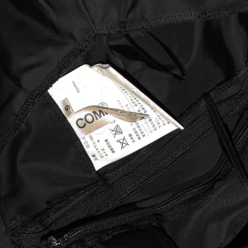 May include: Black clothing label with white text that reads "COMME des GARCONS" and "S" for size. The label also includes Japanese text and washing instructions.