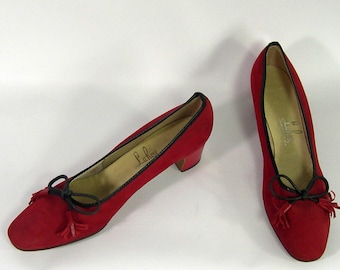 May include: A pair of red suede shoes with black trim and small heels. Each shoe has a black bow with tassel accents. The interior of the shoe has the brand name "La Rose" in script.
