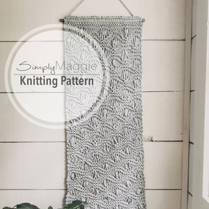 May include: A gray knitted wall hanging with a textured pattern. The hanging is attached to a wooden dowel rod and is displayed against a white wall. The text "Simply Maggie Knitting Pattern" is visible in the image.