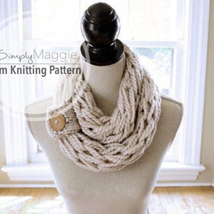 May include: A beige knitted infinity scarf with a wooden button on a white mannequin. The scarf is made using an arm knitting technique. The text "Simply Maggie Arm Knitting Pattern" is visible in the image.