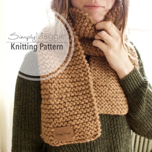 May include: A brown knitted scarf with a simple stitch pattern. The scarf is worn by a person with a green sweater. The scarf has a small leather tag with the text "Simply Maggie" on it.