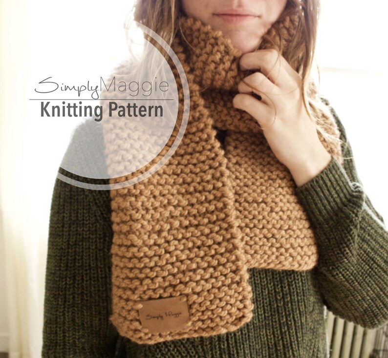 May include: A brown knitted scarf with a simple stitch pattern. The scarf is worn by a person with a green sweater. The scarf has a small leather tag with the text "Simply Maggie" on it.