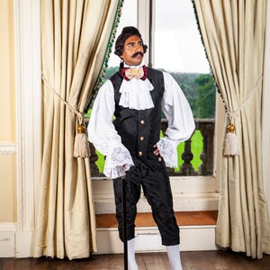 May include: A man wearing a black and white patterned vest with a white shirt and a gold bow tie. He is holding a black walking stick and is standing in front of a window with white curtains.