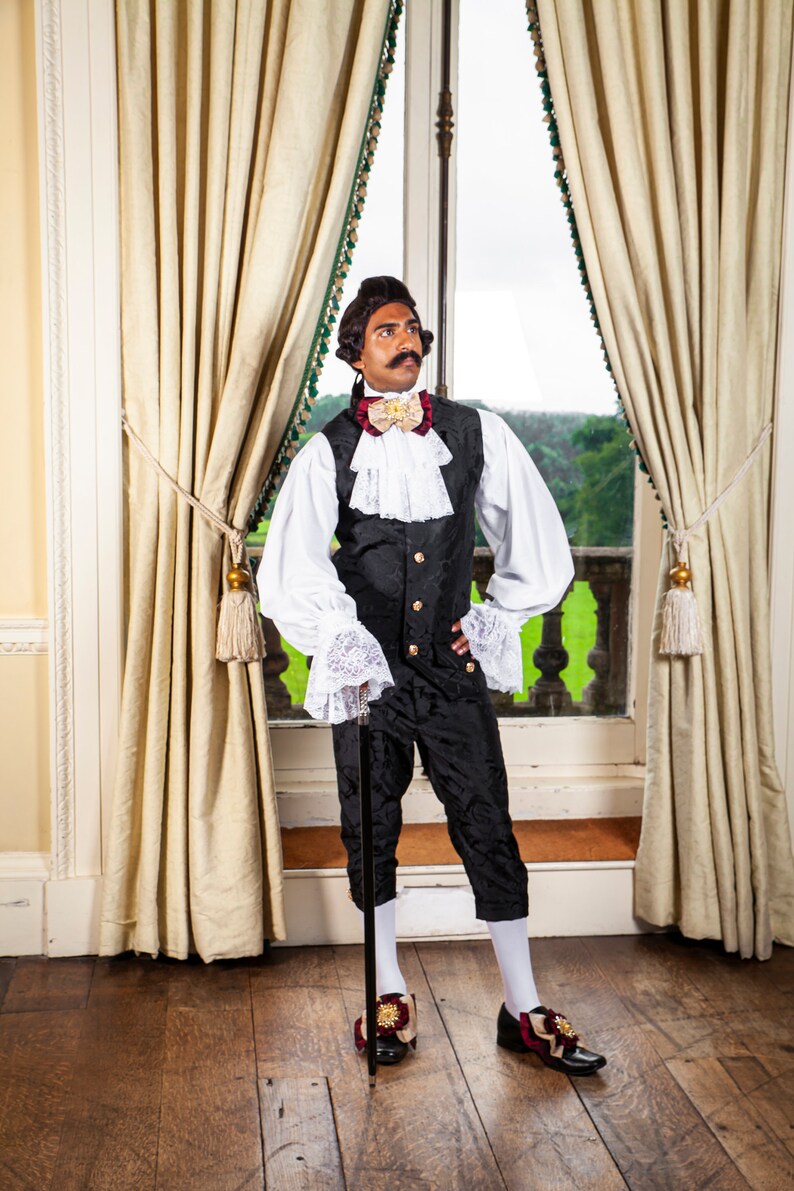 May include: A man wearing a black and white patterned vest with a white shirt and a gold bow tie. He is holding a black walking stick and is standing in front of a window with white curtains.