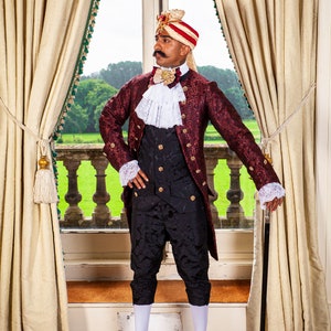May include: A man wearing a red and gold brocade jacket with a white lace collar and a black waistcoat. He is also wearing a red and white turban and black pants. He is holding a black walking stick and standing in front of a window.