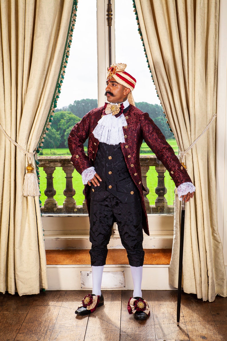 May include: A man wearing a red and gold brocade jacket with a white lace collar and a black waistcoat. He is also wearing a red and white turban and black pants. He is holding a black walking stick and standing in front of a window.