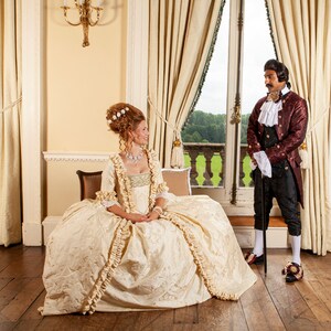 May include: A woman in a white and gold gown with a ruffled skirt stands in a room with a man in a red and gold jacket. The man is holding a cane and the woman is sitting on a couch. The room has large windows with white curtains and a chandelier.