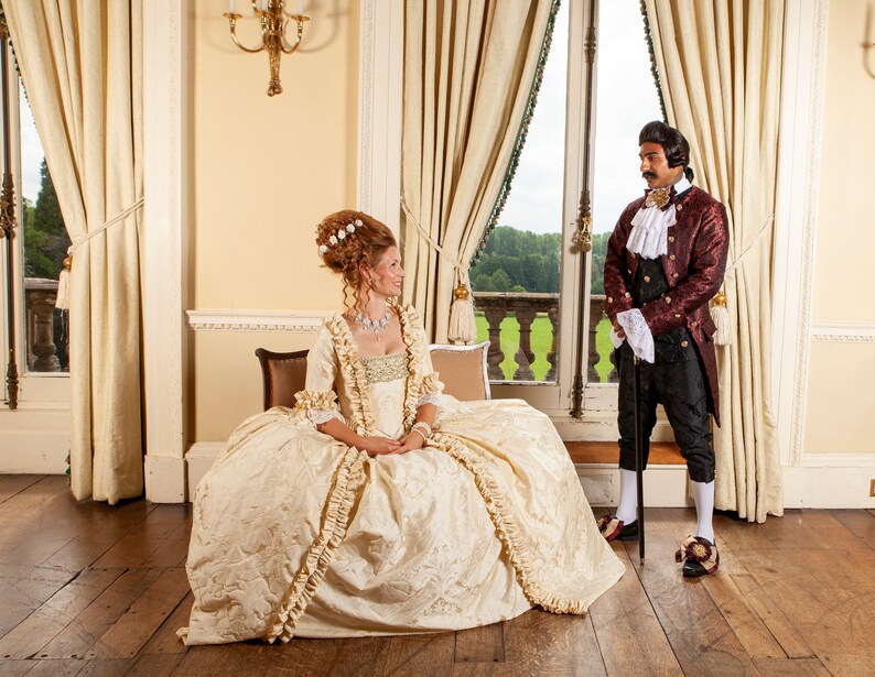 May include: A woman in a white and gold gown with a ruffled skirt stands in a room with a man in a red and gold jacket. The man is holding a cane and the woman is sitting on a couch. The room has large windows with white curtains and a chandelier.