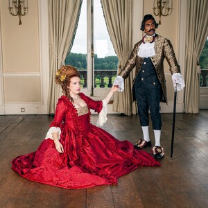 May include: A woman in a red gown with gold accents sits on a wooden floor, reaching out her hand to a man in a gold and black patterned jacket. He is holding a cane and looking away from the camera.