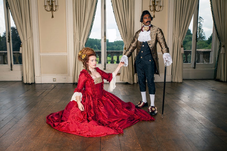 May include: A woman in a red gown with gold accents sits on a wooden floor, reaching out her hand to a man in a gold and black patterned jacket. He is holding a cane and looking away from the camera.