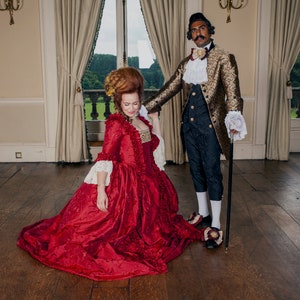 May include: A couple dressed in historical attire. The woman is wearing a long, red gown with lace trim. The man is wearing a gold and black patterned jacket with a white cravat. They are standing in a large room with high ceilings and a wooden floor.