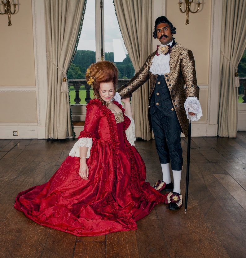 May include: A couple dressed in historical attire. The woman is wearing a long, red gown with lace trim. The man is wearing a gold and black patterned jacket with a white cravat. They are standing in a large room with high ceilings and a wooden floor.