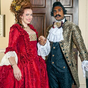 May include: A woman in a red and gold dress with a large, elaborate hairstyle stands next to a man in a gold and black patterned coat. He is wearing a white lace cravat and a gold bow tie. They are both smiling and looking at the camera.