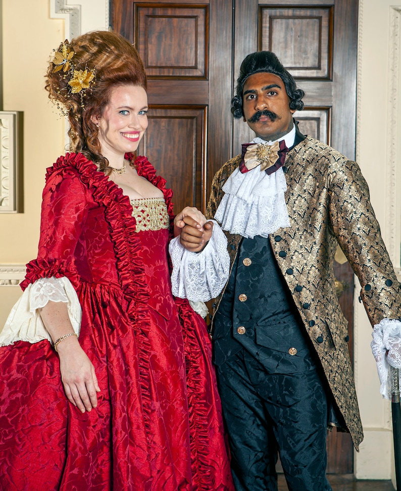 May include: A woman in a red and gold dress with a large, elaborate hairstyle stands next to a man in a gold and black patterned coat. He is wearing a white lace cravat and a gold bow tie. They are both smiling and looking at the camera.