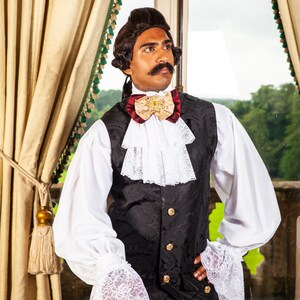 May include: A man wearing a white shirt with lace cuffs, a black vest with gold buttons, and a burgundy and beige bow tie with a gold brooch. He is standing in front of a window with a view of trees.