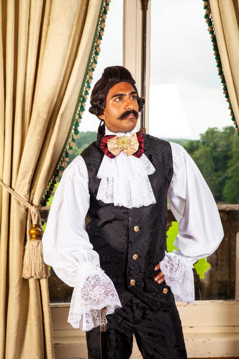May include: A man wearing a white shirt with lace cuffs, a black vest with gold buttons, and a burgundy and beige bow tie with a gold brooch. He is standing in front of a window with a view of trees.