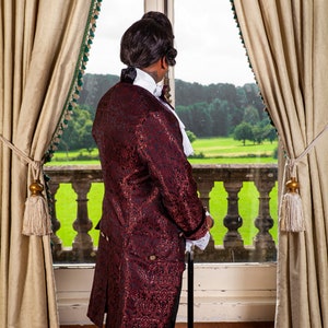 May include: A man in a burgundy brocade coat with black trim stands by a window looking out at a green lawn. He is wearing a white shirt with a lace collar and black pants. He is holding a black walking stick.