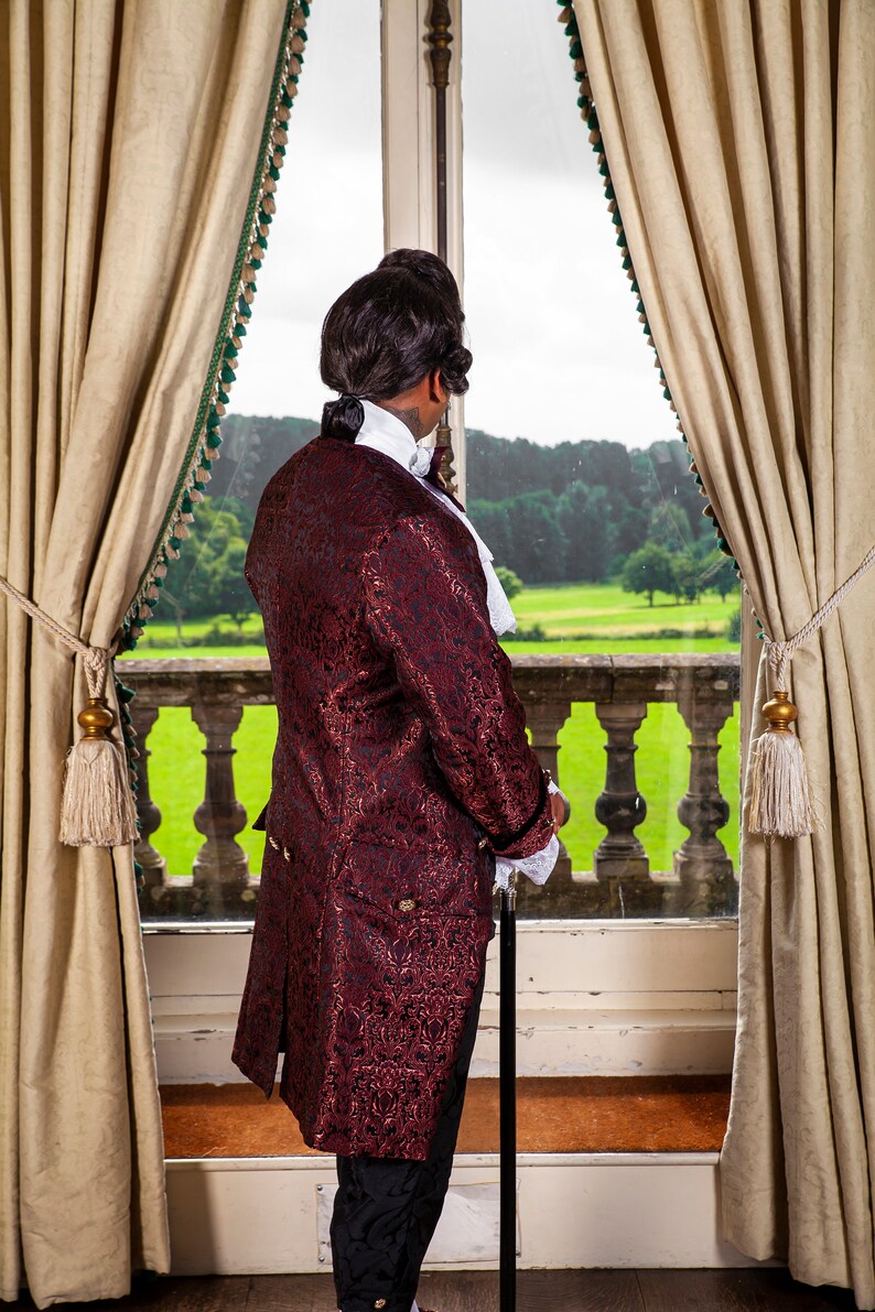 May include: A man in a burgundy brocade coat with black trim stands by a window looking out at a green lawn. He is wearing a white shirt with a lace collar and black pants. He is holding a black walking stick.