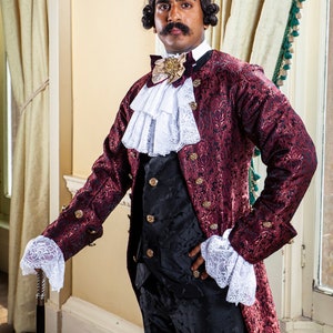 May include: A man wearing a burgundy brocade jacket with gold accents, a white lace cravat, and black pants. He is holding a black walking stick.