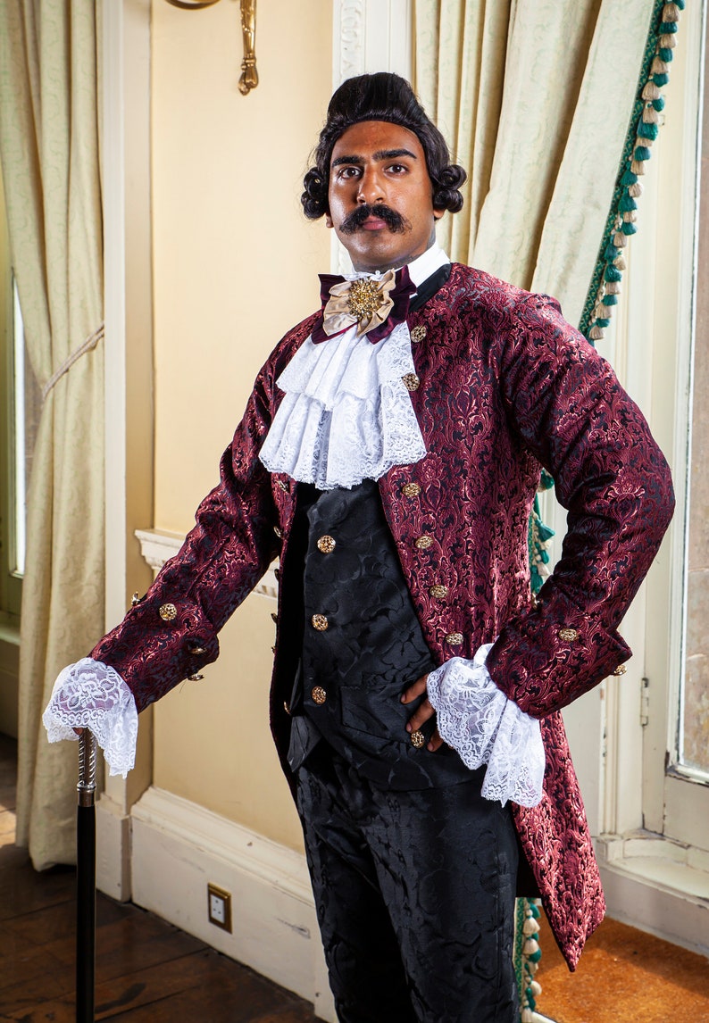 May include: A man wearing a burgundy brocade jacket with gold accents, a white lace cravat, and black pants. He is holding a black walking stick.