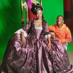 May include: A woman in a purple gown with a large skirt and a white lace bodice sits on a gold throne. She is wearing a pearl and feather headpiece. A man in an orange hoodie stands behind her.