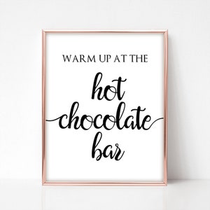 May include: A framed print with a rose gold border and a white background. The black text reads "WARM UP AT THE hot chocolate bar" in a mix of fonts. The print is a decorative item.