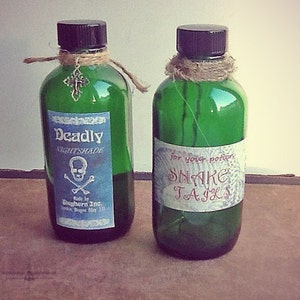 K&ouml;nnte beinhalten: Zwei gr&uuml;ne Glasflaschen mit schwarzen Deckeln, beschriftet mit "Deadly Nightshade" und "Snake Tails". Die "Deadly Nightshade"-Flasche hat einen Kreuzanh&auml;nger und Bindfaden. F&uuml;r Zaubertr&auml;nke.