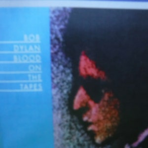 May include: Album cover with the text "BOB DYLAN BLOOD ON THE TAPES" in white on a blue background. A blurred image of a person's face is visible on the right side of the cover, with red and orange tones.