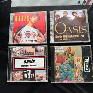 May include: Four Oasis CD cases with different album covers. The top left case features a red background with the band's name in white and the album title "Due North" in white. The top right case features a black background with the band's name in gold and the album title "Live On Swedish Radio '94 and More..." in white. The bottom left case features a collage of images with the band's name in white and the album title "Backbeat: Volume 2" in white. The bottom right case features a comic book style image with the band's name in white.